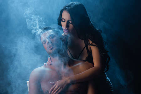 Brunette woman touching sexy man on black background with smokeの写真素材