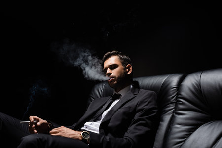 Brunette man in formal wear smoking on leather couch isolated on blackの写真素材