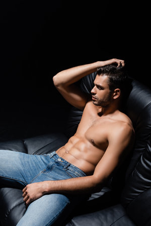 Shirtless man in jeans sitting on leather couch isolated on blackの写真素材