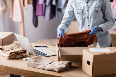 cropped view of seller in latex gloves packing clothes into boxes in fashion boutique