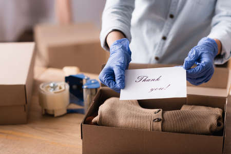 partial view of fashion boutique seller in latex gloves holding card with thank you lettering, blurred backgroundの写真素材