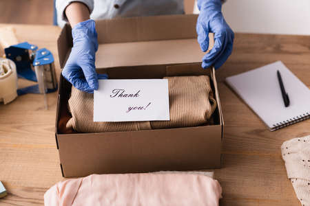 cropped view of showroom owner in latex gloves packing card with thank you inscription into box, blurred backgroundの写真素材