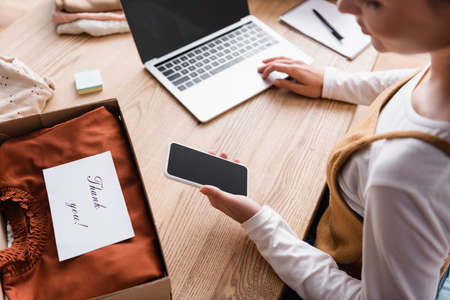 partial view of showroom owner holding smartphone with blank screen at workplace, blurred foreground
