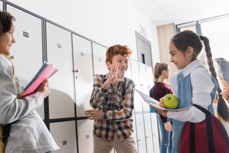 Positive schoolchild talking with schoolgirl holding apple and copybook on blurred foreground in schoolの写真素材