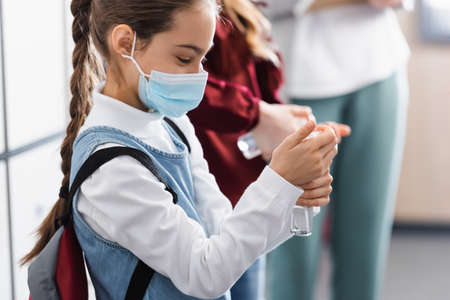 Schoolgirl in medical mask using hand sanitizer near classmate and teacher on blurred backgroundの写真素材
