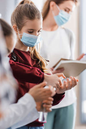 Pupil in protective mask using hand sanitizer near friend and teacher on blurred backgroundの写真素材