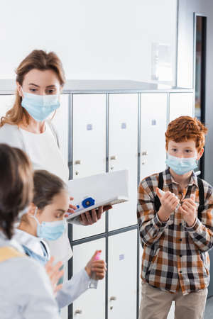 Boy in medical mask holding hand sanitizer near teacher with infrared thermometer and notebook in corridorの写真素材