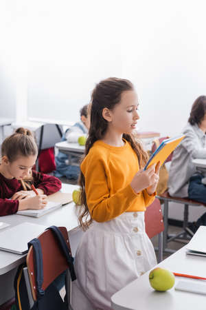 Schoolgirl reading from notebook near devices, apple and classmates on blurred background in classroomの写真素材
