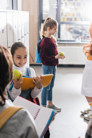 Cheerful schoolchild with apple and notebook looking at classmates on blurred foreground in hallの写真素材