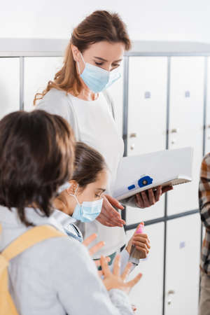 Teacher with notebook and infrared thermometer standing near pupils in protective masks with disinfectorの写真素材
