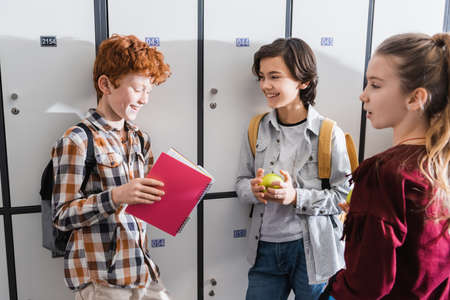 Cheerful kids with notebook, apples and backpacks standing near lockers in schoolの写真素材
