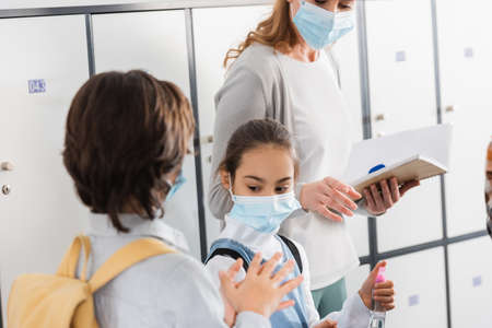 Schoolgirl holding disinfector near friend and teacher with notebook on blurred backgroundの写真素材