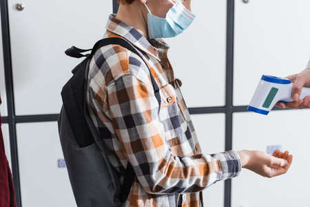 Cropped view of schoolboy in medical mask standing near teacher measuring temperature with infrared thermometer, bannerの写真素材