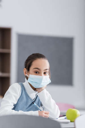 Schoolkid in medical mask holding pen and looking at camera in classroom
