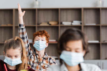 redhead schoolboy in medical mask raising hand during lesson near classmates on blurred foreground