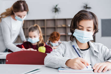 schoolboy in medical mask writing in notebook near classmates and teacher on blurred background