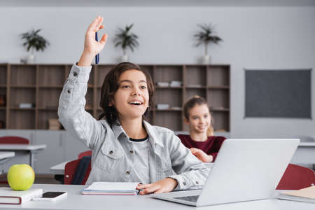 Cheerful boy raising hand near notebook and devices in classroom