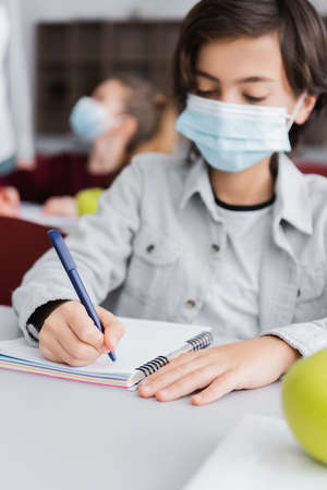schoolboy in medical mask writing in notebook on blurred backgroundの写真素材