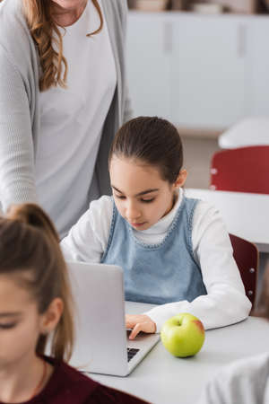 schoolgirl working on laptop near apple and teacher standing nearby