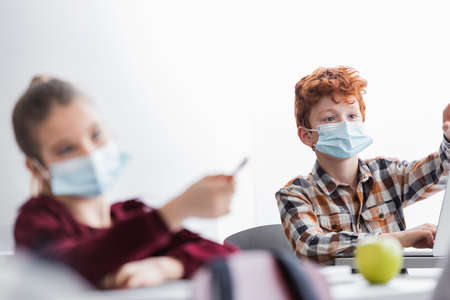redhead schoolboy in medical mask near girl pointing with pen on blurred foreground
