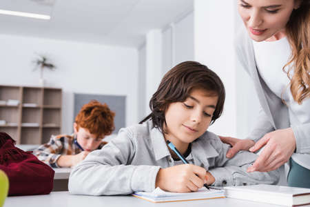 schoolboy writing in notebook near teacher helping him during lessonの写真素材
