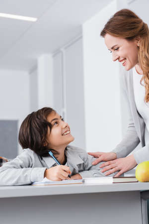 happy teacher and schoolboy looking at each other during lesson in schoolの写真素材
