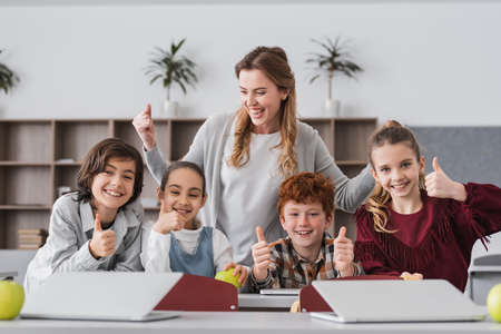 excited schoolkids and teacher showing thumbs up in classroom