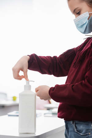 side view of schoolgirl in medical mask applying antiseptic on hands in classroomの写真素材