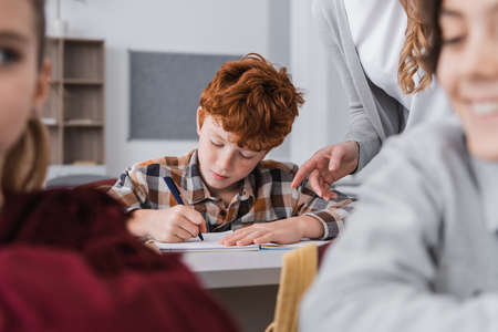 teacher pointing with finger near redhead boy writing during lesson near pupils on blurred...