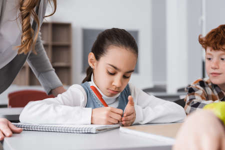 schoolgirl writing in notebook near teacher and classmates on blurred foreground