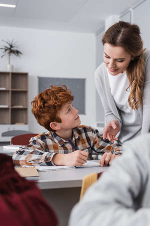 smiling teacher helping redhead kid during lesson, blurred foregroundの写真素材
