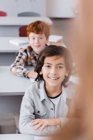 cheerful boy looking at camera in classroom on blurred foreground