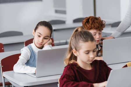 bored schoolgirl sitting near laptop during lesson near classmates on blurred foregroundの写真素材