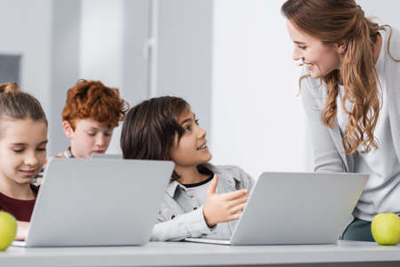 schoolboy pointing at laptop near smiling teacher in classroom