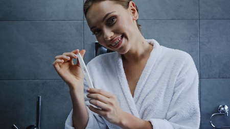 cheerful young woman in bathrobe holding pregnancy test in bathroomの写真素材