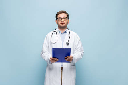 doctor in white coat holding clipboard and looking at camera on blueの写真素材