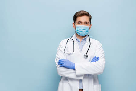 doctor in medical mask and white coat with stethoscope standing with crossed arms isolated on blueの写真素材