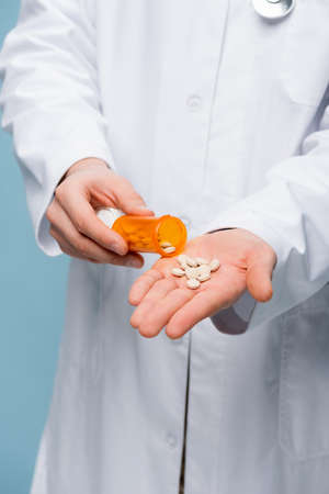 partial view of doctor in white coat holding pills and bottle in hands isolated on blueの写真素材