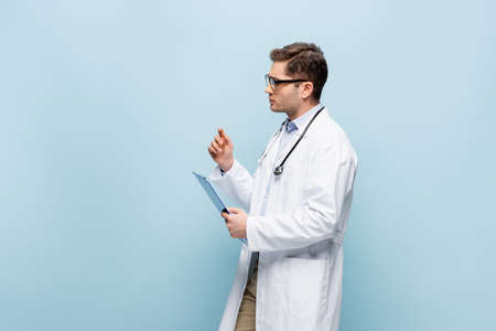 young doctor in glasses and white coat holding clipboard isolated on blue