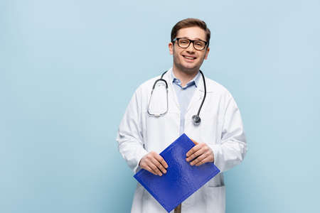 smiling doctor in glasses and white coat holding clipboard and looking at camera on blue