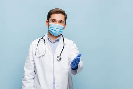 doctor in medical mask and white coat with stethoscope pointing with hand isolated on blueの写真素材