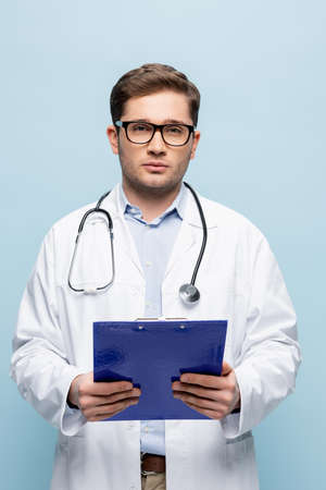 young doctor in glasses and white coat holding clipboard and looking at camera isolated on blue
