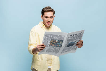 surprised young man in shirt reading travel newspaper isolated on blueの写真素材