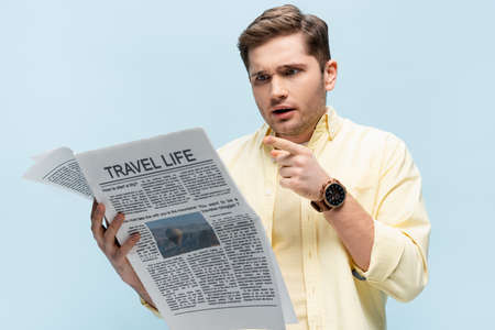 young man in shirt pointing with finger at travel newspaper isolated on blueの写真素材