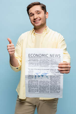 happy young man in shirt holding economical newspaper and showing thumb up isolated on blue