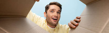 bottom view of curious young man looking at camera from carton box isolated on blue, banner