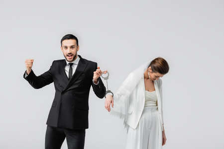 Excited muslim groom holding handcuffs of upset bride isolated on gray