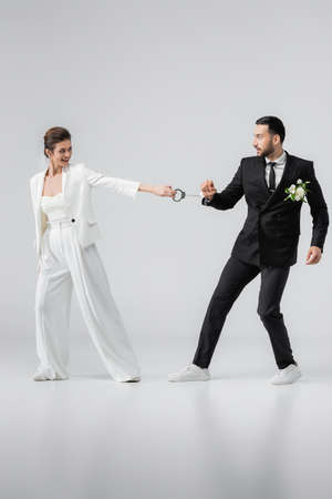 Cheerful bride holding handcuffs of arabian groom on white background
