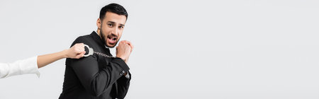 Scared arabian groom in handcuffs looking at camera near bride isolated on gray, bannerの写真素材