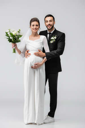 Happy arabian groom hugging pregnant bride and looking at camera on gray backgroundの写真素材